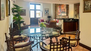 A dining area with a glass table surrounded by wooden chairs, a large potted plant, and a view of the kitchen in the background.