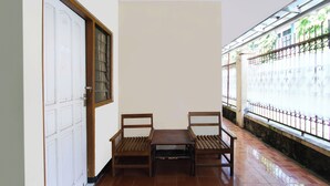 Ein Zimmer mit Fliesenboden, zwei Holzstühlen, einem kleinen Tisch, einer weißen Tür mit Fensterteil und Blick auf einen Garten durch einen Metallzaun.