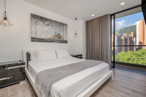 Quarto moderno com uma cama grande, mesa de cabeceira, pintura e vista para edifícios e áreas verdes.