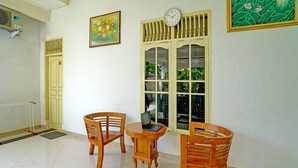 Ein Zimmer mit Holzstuhl, einem kleinen runden Tisch mit einem Laptop, einer Wanduhr und einem Fenster mit Blick ins Grüne.