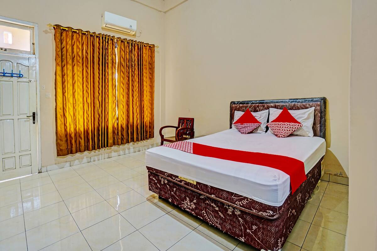 Una habitación de hotel con cama, una silla, ventana con cortinas y unidad de aire acondicionado.