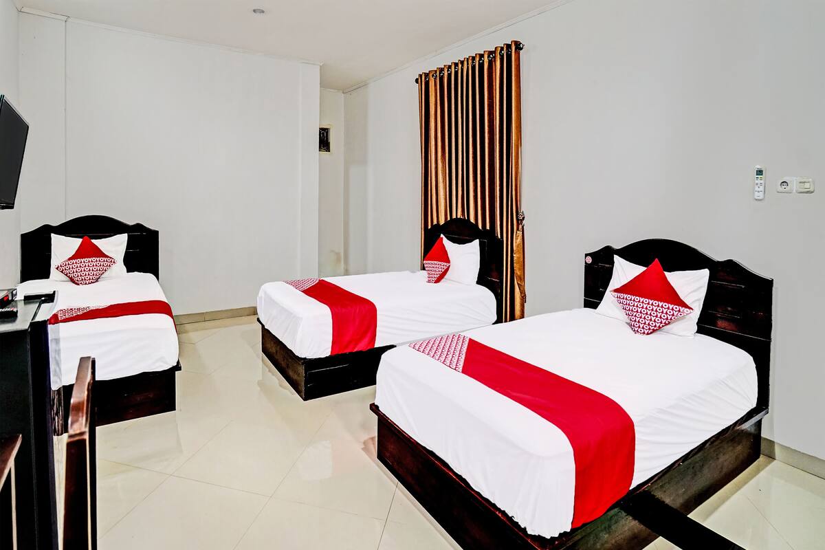 Habitación de hotel con tres camas, cada una con ropa de cama blanca y detalles rojos, cabeceros de madera y un ventanal grande con persianas verticales.