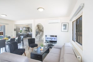 A modern living room with a sofa, coffee table, and TV. There is a dining area with chairs and a table. A kitchen is visible in the background.
