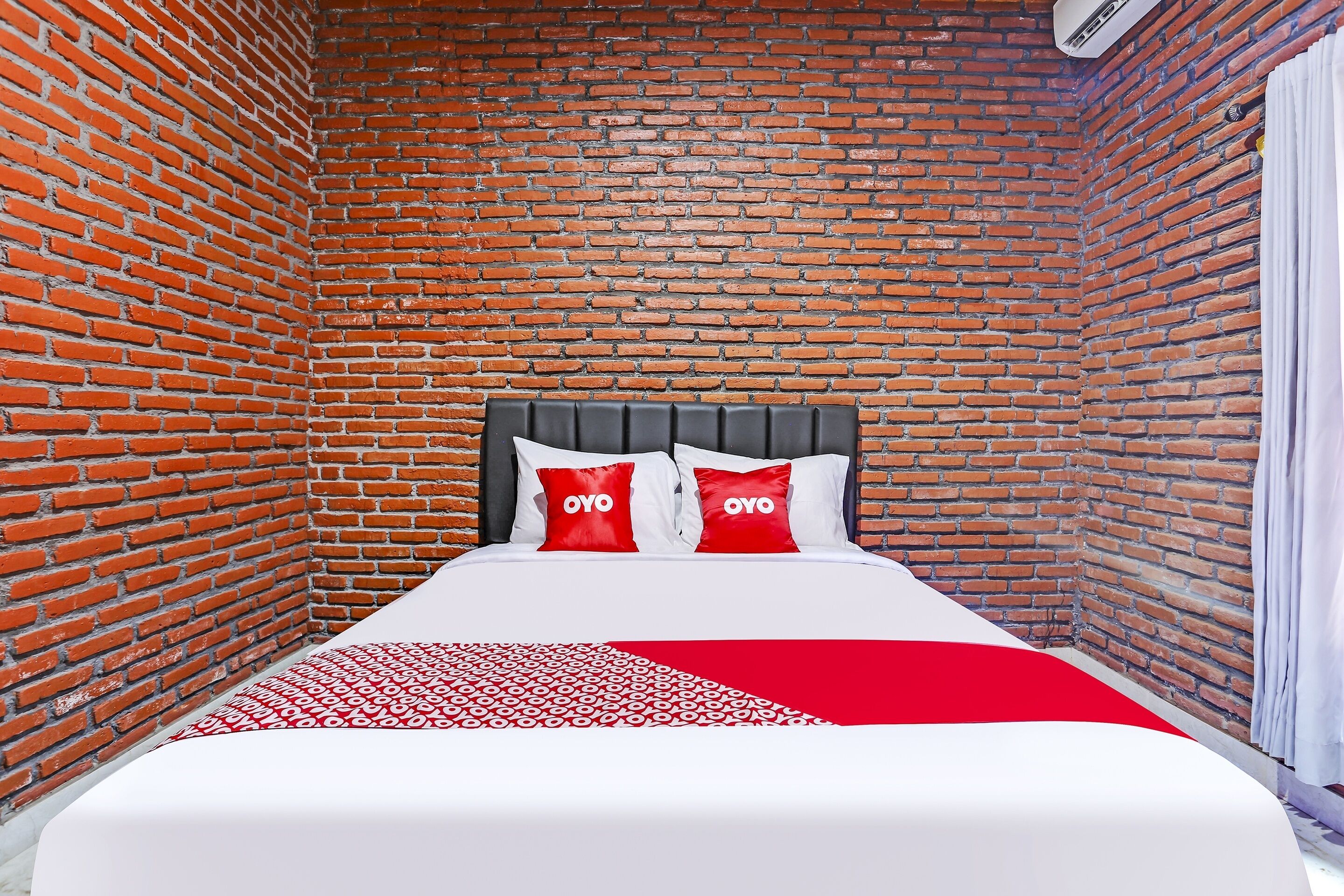 A room with a brick wall, a bed with white bedding and red pillows, and a red and white patterned throw.
