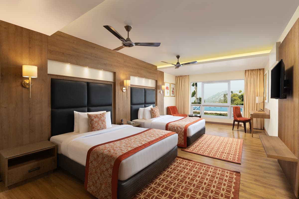 A hotel room with two beds, a desk, a chair, a TV, and a balcony with a view.