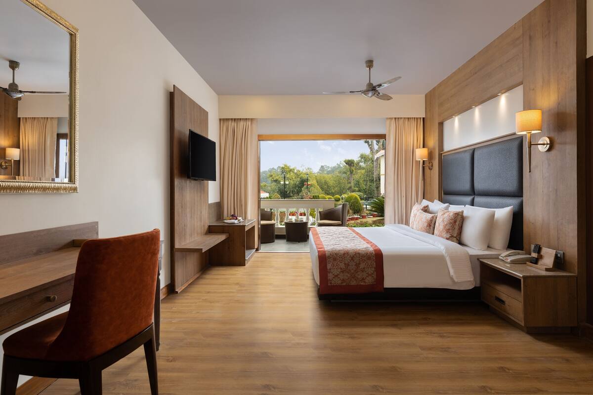 A hotel room with a bed, a desk, a chair, a TV, and a balcony with a view of greenery.