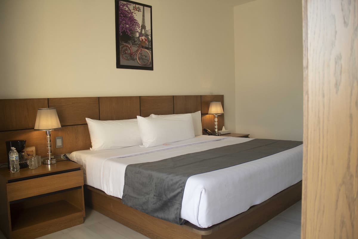 Una cama impecablemente arreglada con sábanas blancas y un edredón gris, flanqueada por mesitas de noche de madera con lámparas, en una habitación con cabecera de madera y un cuadro enmarcado en la pared.