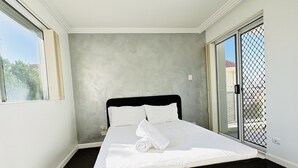A neatly made bed with white linens and a rolled towel, a dark wooden headboard, and a carpeted floor in a room with a large window and a sliding glass door. - Modern Apartment in Chevron Island (Surfers Paradise)