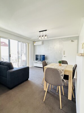 Relax in this spacious living area with a dining table, sofa, and TV, leading out to an outdoor space.