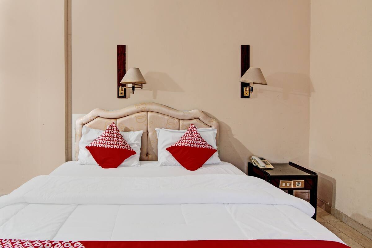 A neatly made bed with white bedding and red pillows, a bedside table with a lamp, and a phone.