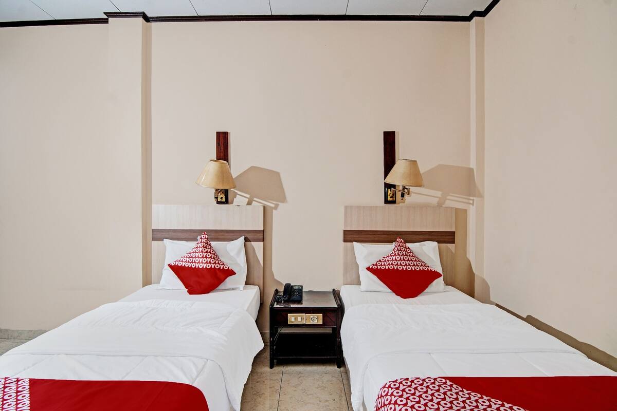 A hotel room with two beds, each with a wooden headboard, white bedding, and red pillows. There are bedside lamps and a small table with a phone between the beds.