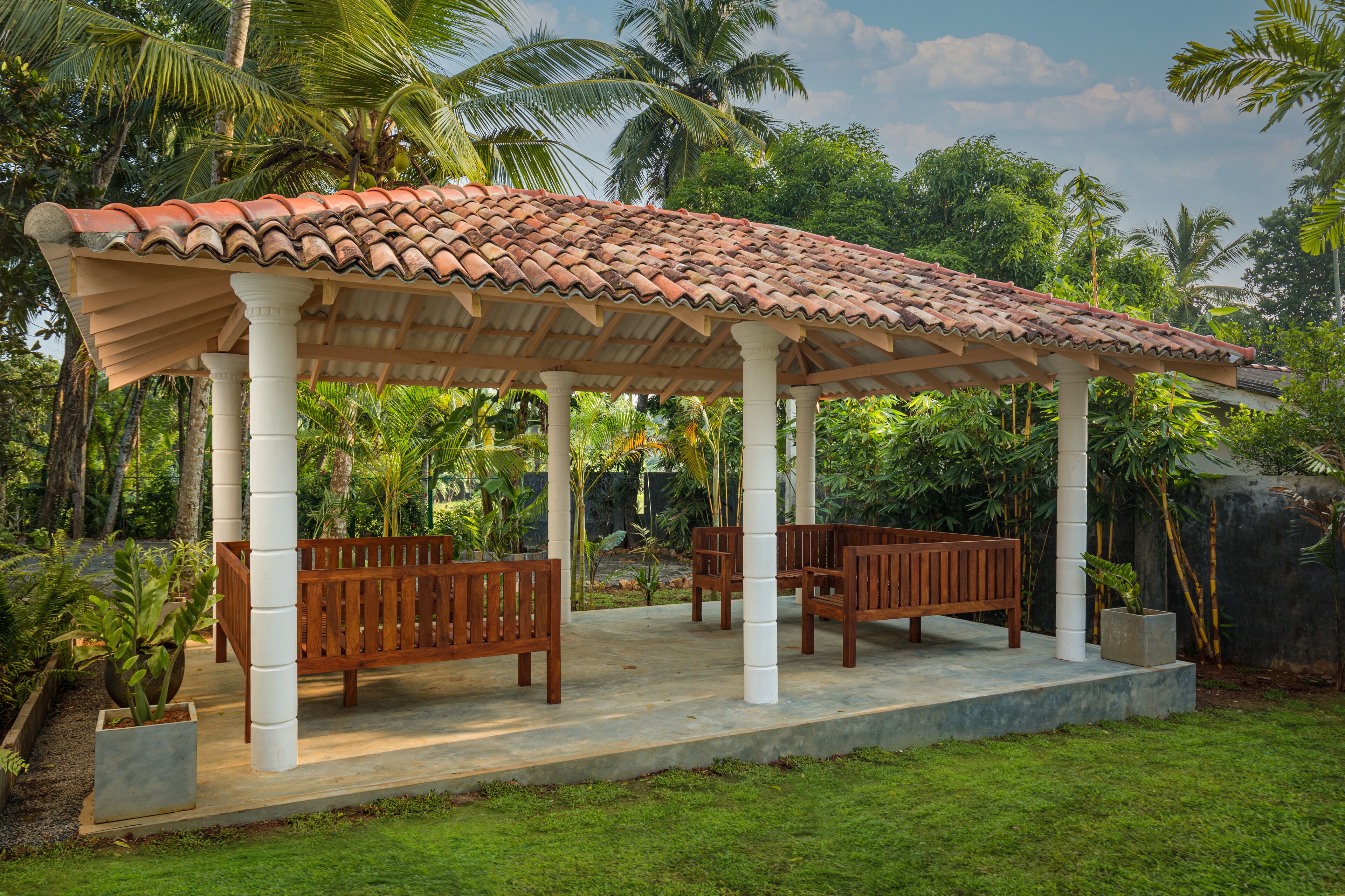 Un área de estar al aire libre techada, con bancos de madera y techo de baldosas, rodeada de exuberante vegetación y palmeras.