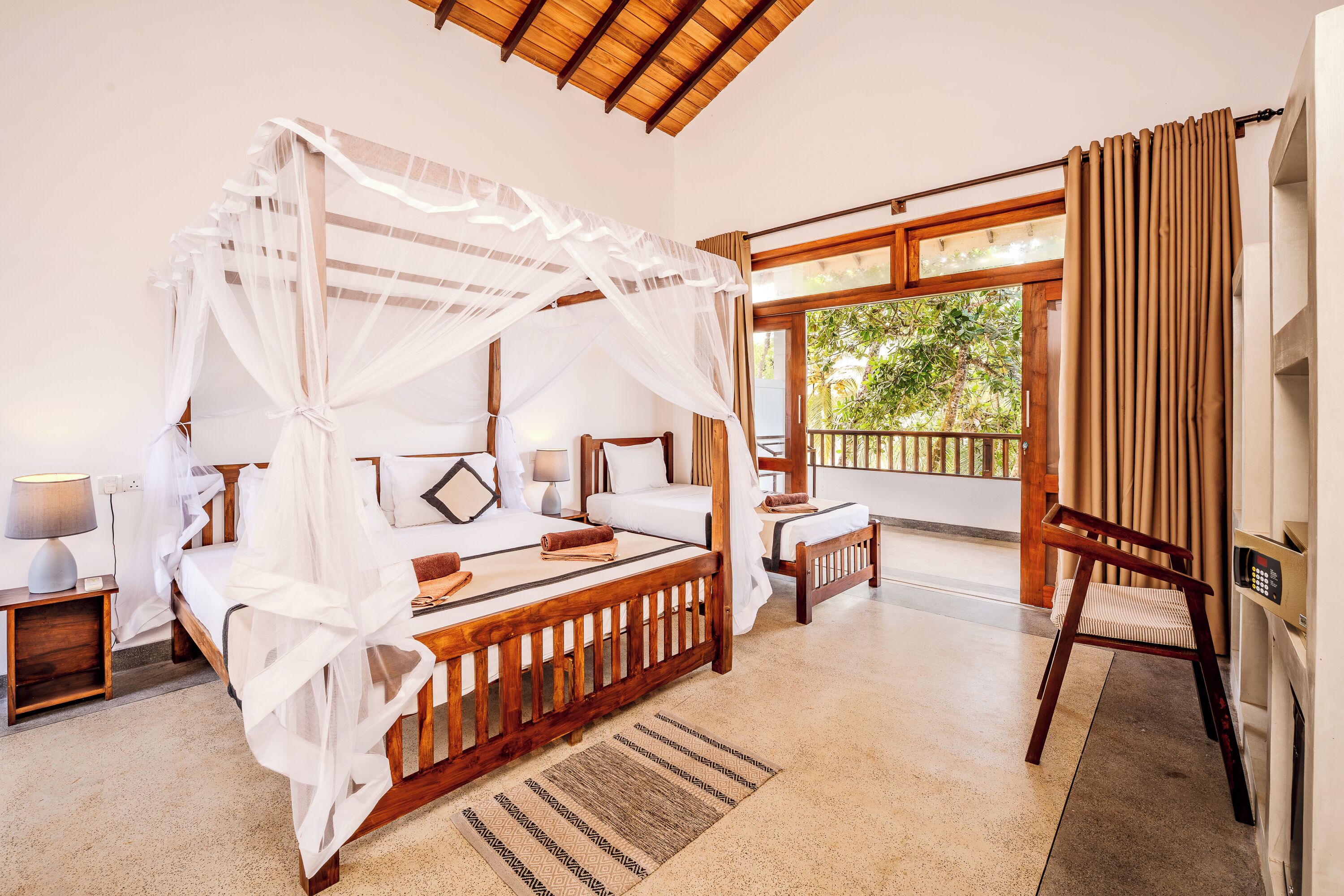 A room with a four-poster bed, a wooden chair, a small table, and a view of greenery outside.