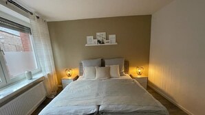 Un dormitorio con una cama grande, dos mesitas de noche con lámparas, una ventana con persianas y un estante en la pared con objetos.
