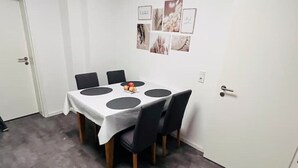 Un comedor con una mesa redonda, seis sillas y un mantel blanco. Se observan adornos en la pared y una puerta al fondo.