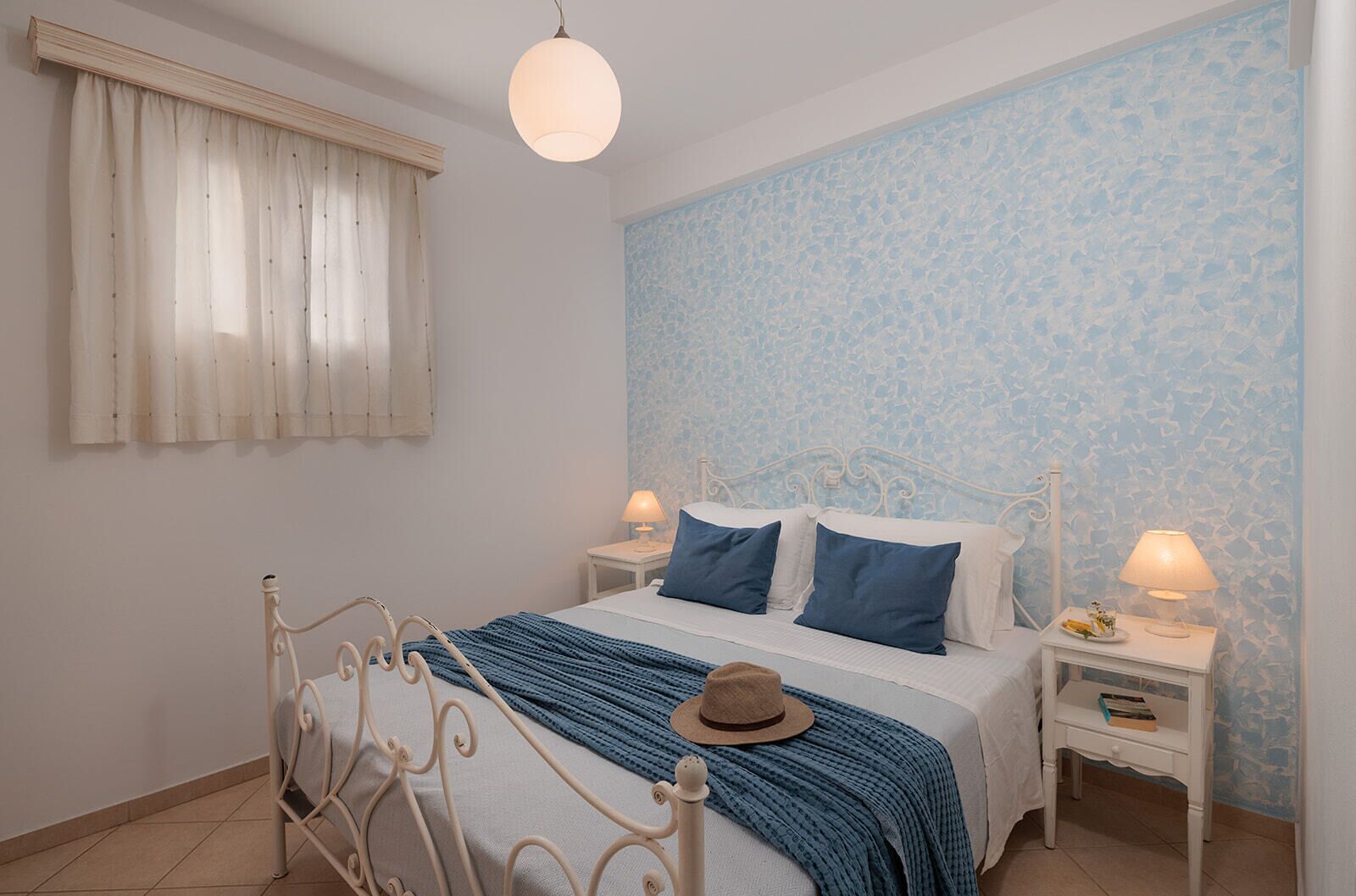 Un dormitorio con una cama, dos mesitas de noche, una lámpara colgante, una ventana con cortinas y una pared estampada.