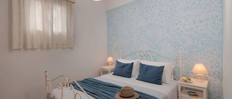 Un dormitorio con una cama, dos mesitas de noche, una lámpara colgante, una ventana con cortinas y una pared estampada.