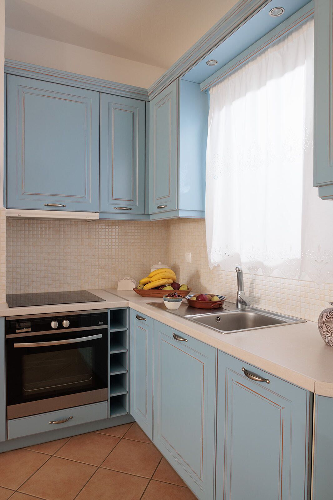 A kitchen with light blue cabinets, a built-in oven, a sink, and a window with a sheer curtain.