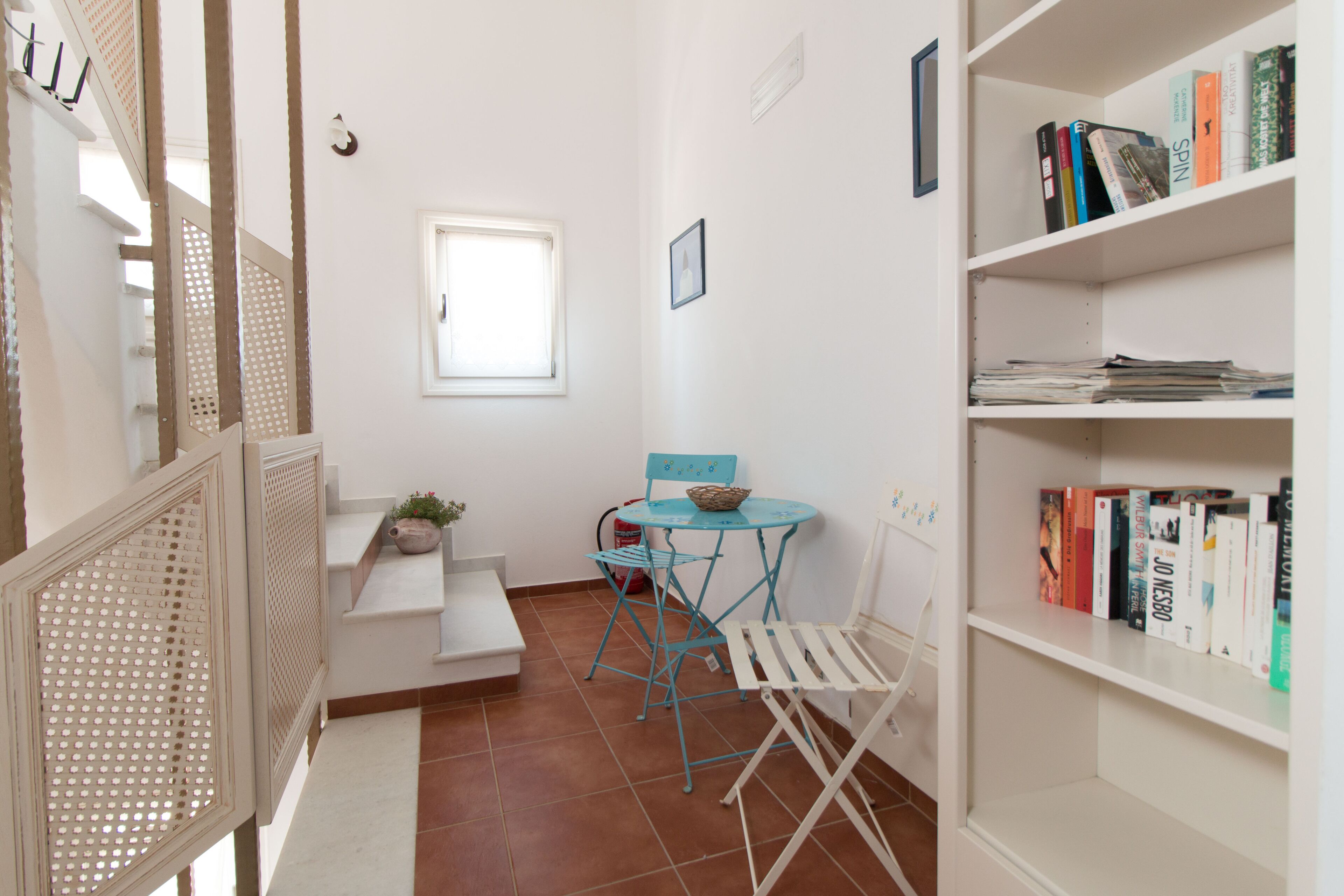 A small, bright room with a window, a blue table with chairs, a bookshelf, and a white radiator.