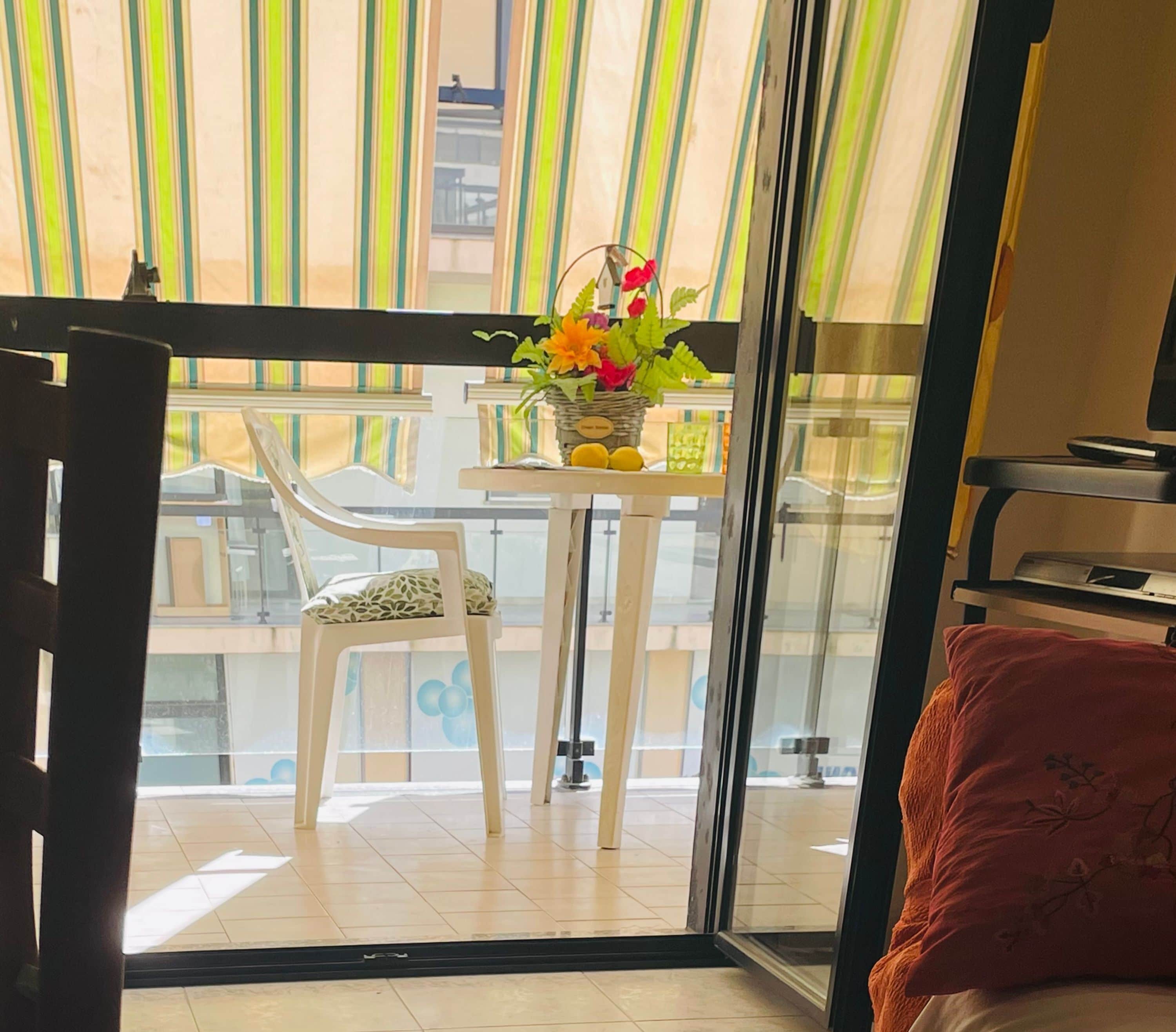 A balcony with a chair, a table with a vase of flowers, and a view of buildings outside.