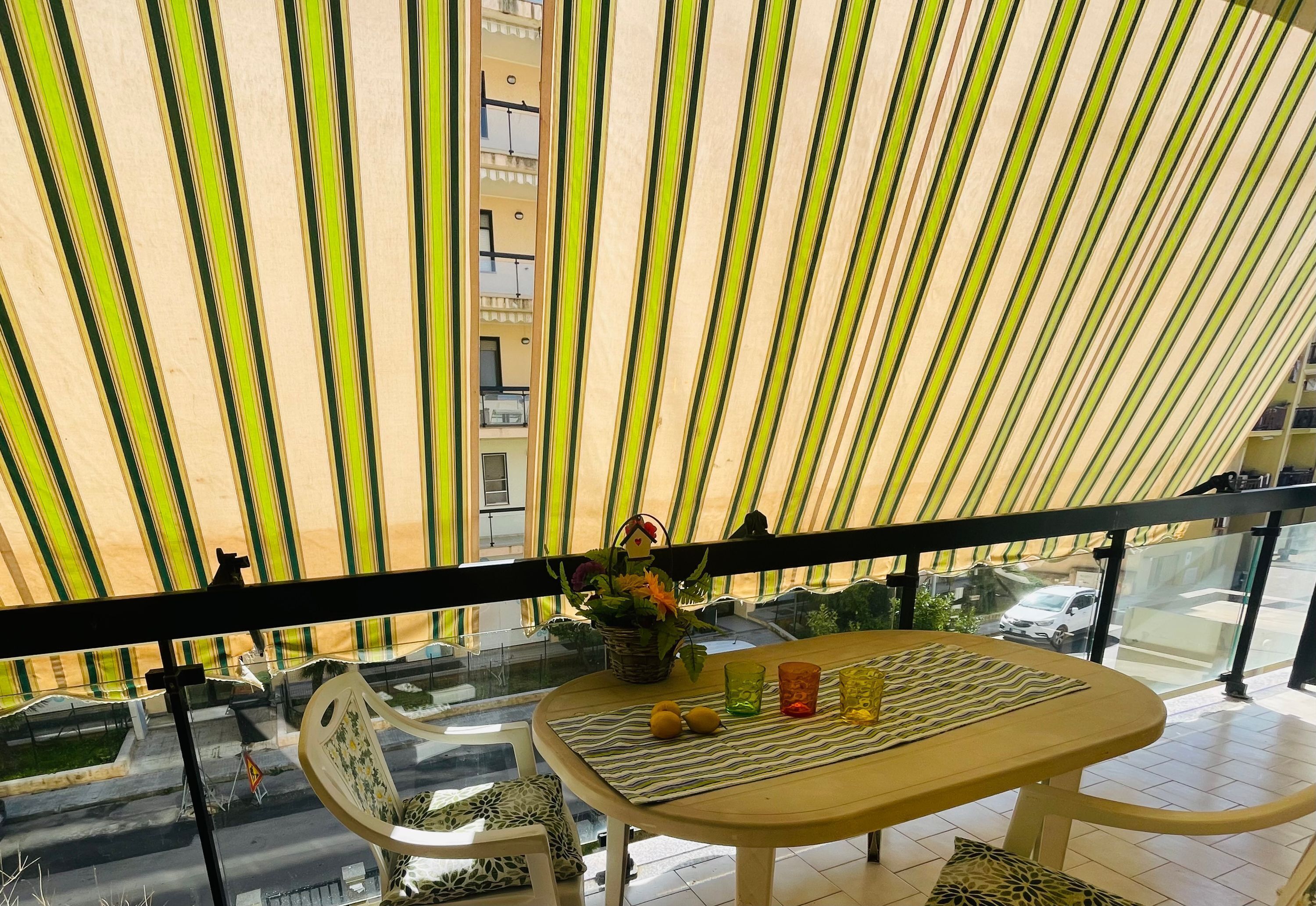 Ein Balkon mit gestreiftem Sonnenschirm, einem Tisch mit Unterlage, einem Blumenkorb und Blick auf die Straße.