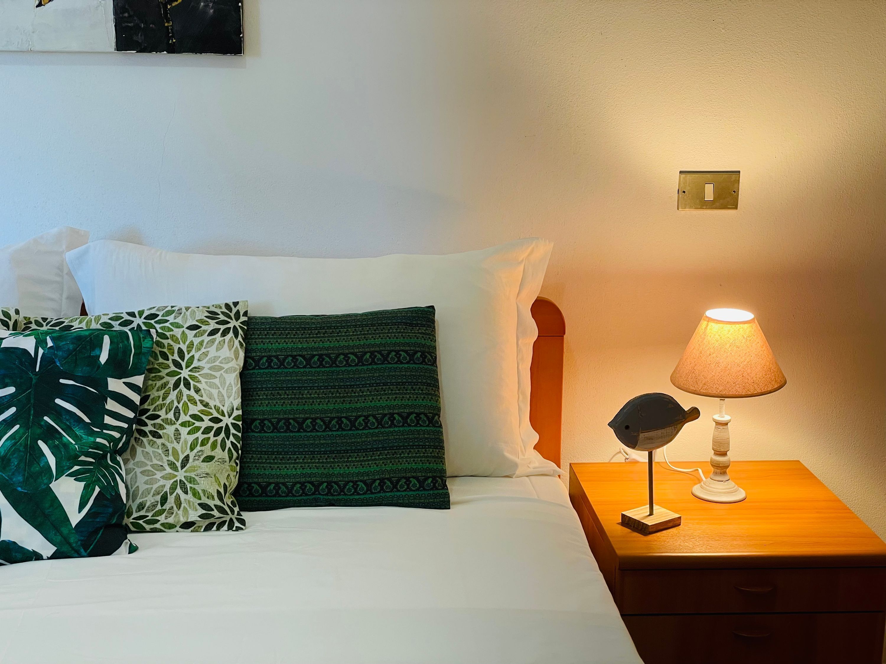 A bed with white and green patterned pillows, a wooden nightstand with a lamp, and a wall switch.