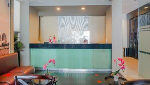 Un espace d’accueil d’hôtel avec un comptoir vert, un canapé en cuir marron, une petite table sur laquelle est posé un vase rempli de fleurs roses, et une cloison en verre.