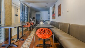 Un espace repas moderne avec de longues tables et des chaises orange, un canapé en cuir et un tapis à motifs.