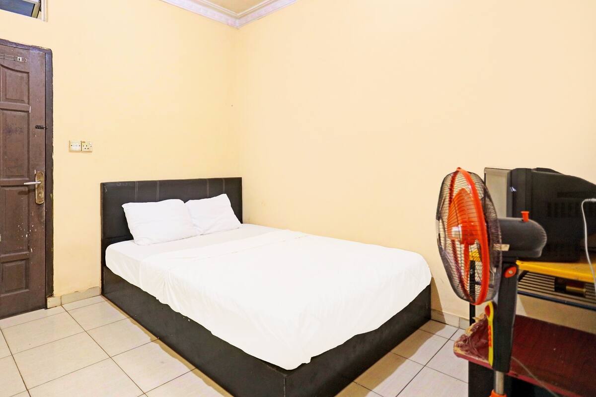 A bedroom with a single bed, a wooden door, a wall socket, and a standing fan.