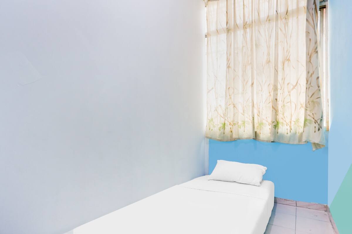 A single bed with a white mattress and pillow, a window with a bamboo-patterned curtain, and a blue wall.