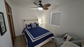 Une chambre à coucher avec un lit, un ventilateur de plafond, une fenêtre avec des stores et une chaise.
