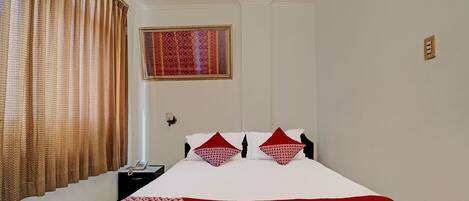 A hotel room with a bed, a nightstand, a telephone, a window with curtains, and a decorative wall hanging.