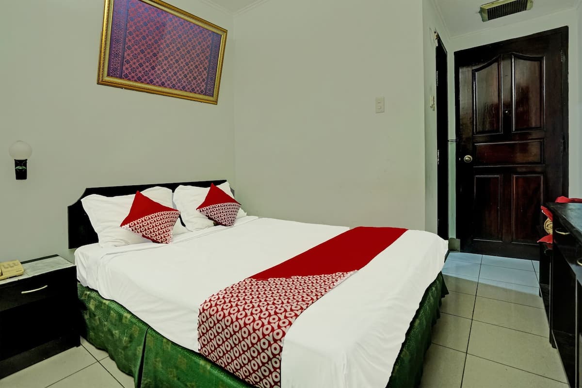 Un lit bien fait, recouvert d’une couvre-lit blanche et rouge à motifs, avec un couvre-lit vert et deux oreillers rouges. Un tableau encadré est accroché au mur au-dessus du lit, une table de chevet avec une lampe se trouve à côté, et une porte en bois est située à droite.