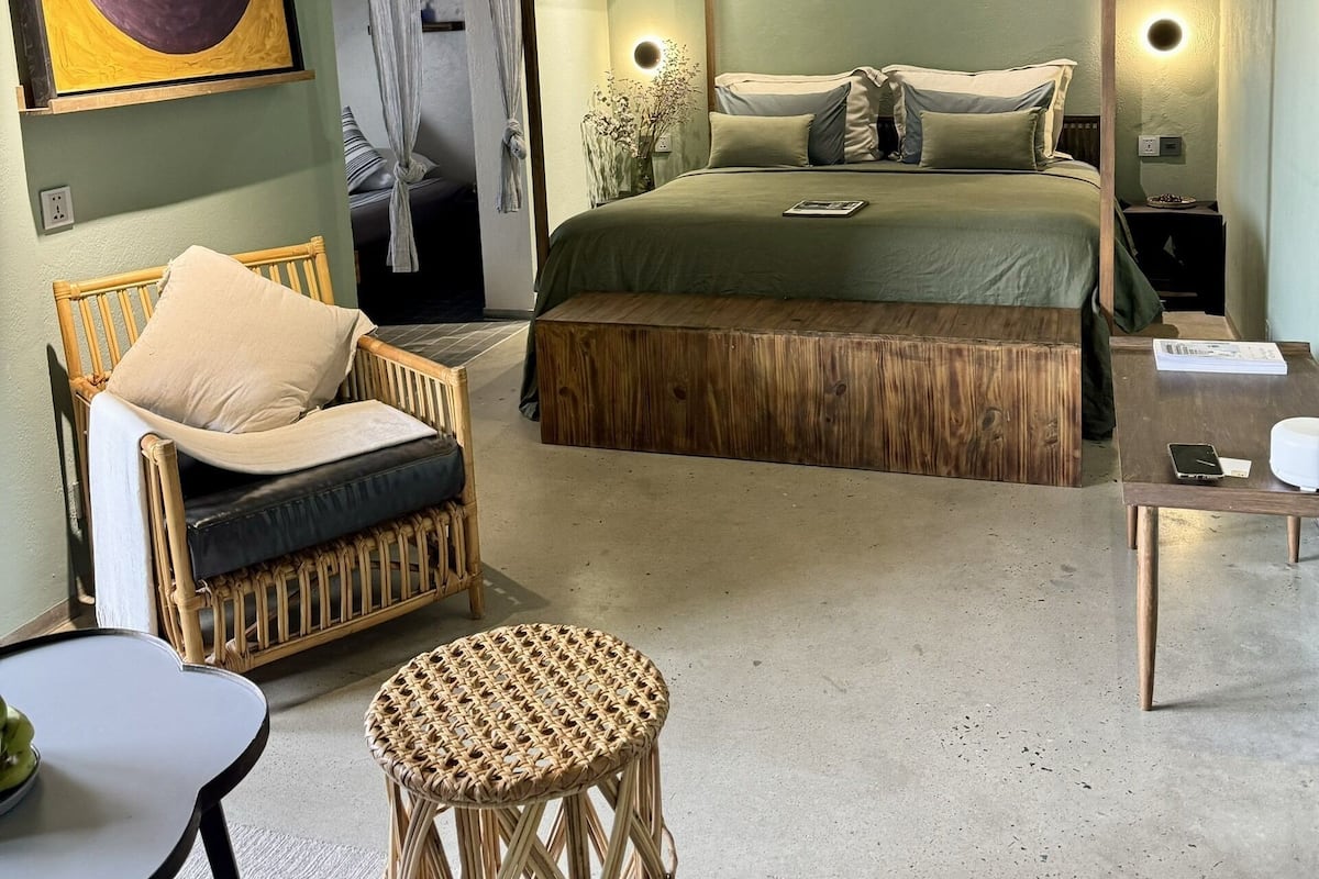 A room with a four-poster bed, a wicker chair, a small round table, and a large abstract painting on the wall.