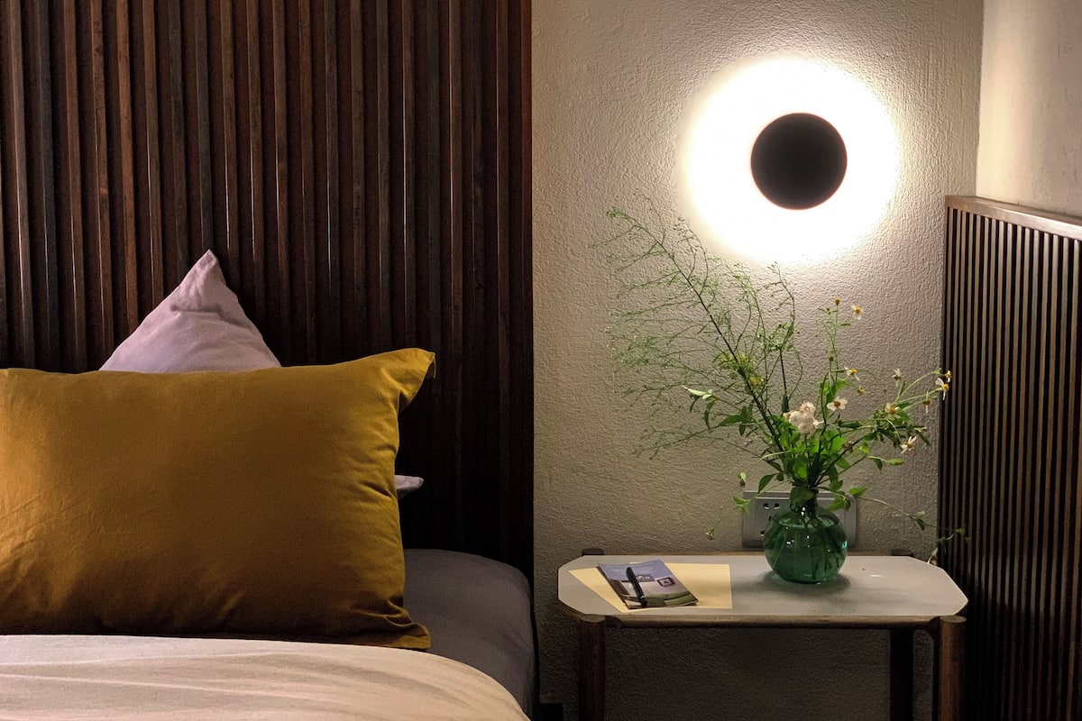 A hotel room with a bed, a bedside table with a vase of flowers, and a wall-mounted light fixture.