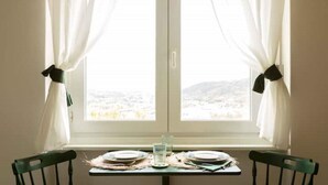 Una mesa de comedor con platos y un vaso, frente a una ventana con cortinas blancas recogidas, mostrando una vista del paisaje.
