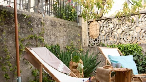 Una terraza de madera con una silla de descanso, una mesa pequeña con una cesta y una taza, y una cesta de mimbre colgada en una valla metálica.