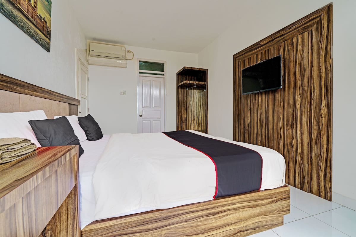 Un dormitorio con cabecera de madera, cama con ropa blanca y un detalle rojo, un televisor de pantalla plana montado en una pared de madera, y una puerta con marco verde.