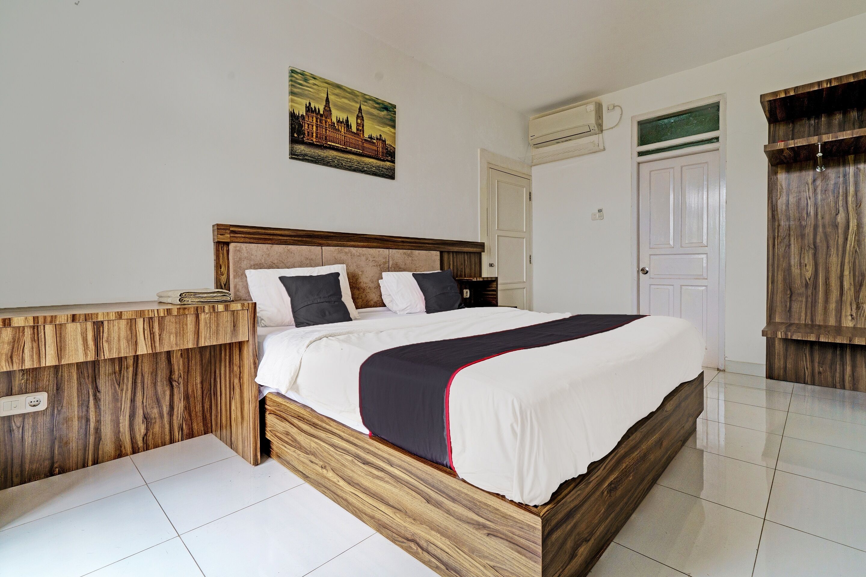 A bedroom with a wooden bed frame, white bedding, and a cityscape painting on the wall.
