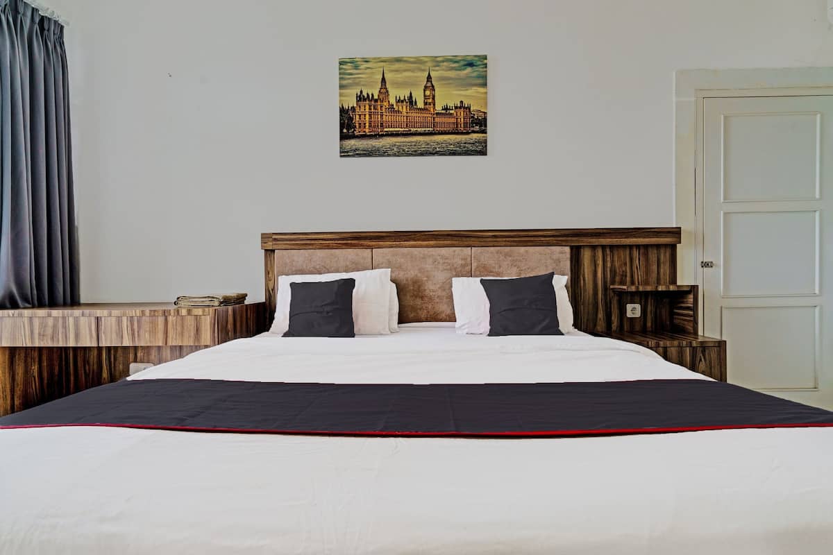 Una cama bien hecha con cabecera de madera, una mesita de noche con lámpara y un cuadro enmarcado de un paisaje urbano en la pared.
