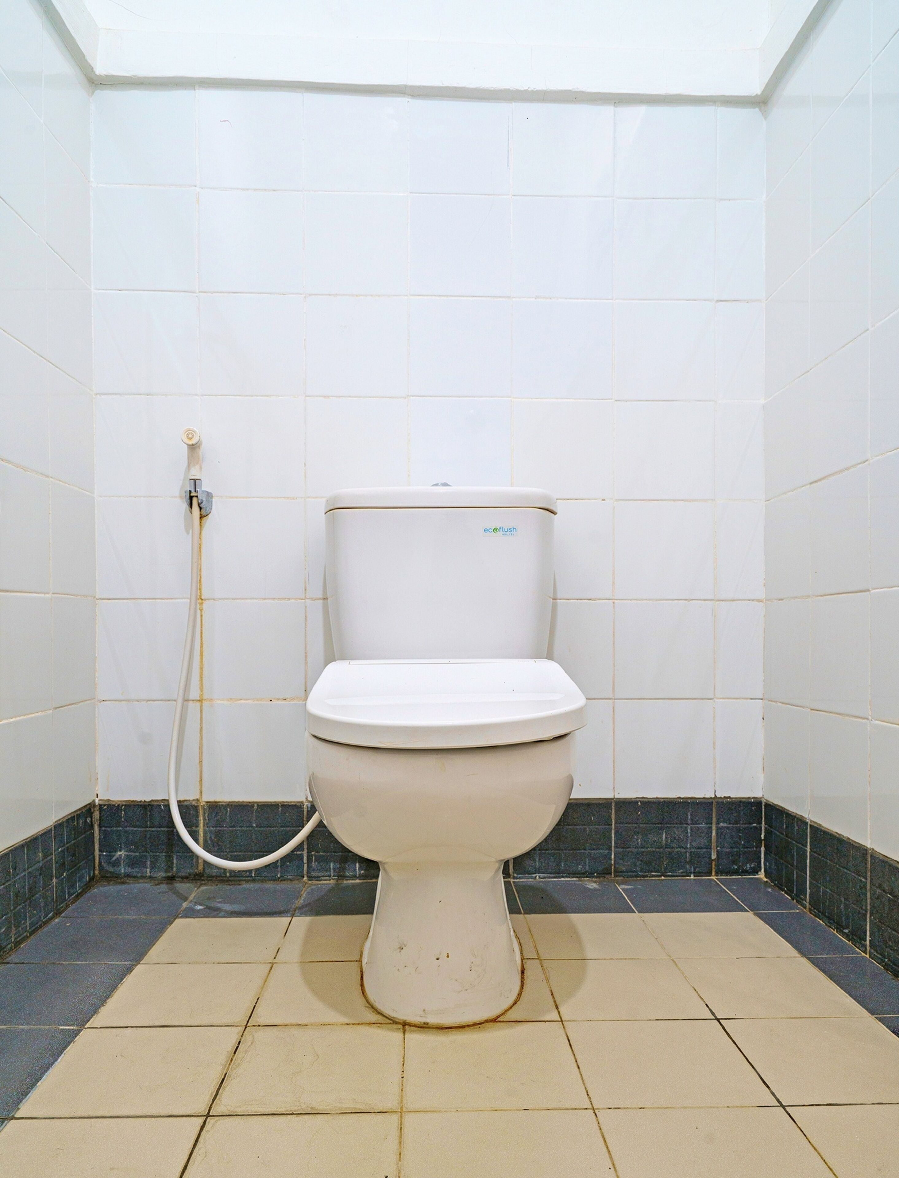 Des toilettes blanches classiques, dotées d’une poignée de chasse d’eau et d’un tuyau d’alimentation en eau, dans une salle de bain carrelée avec un espace douche d’angle.
