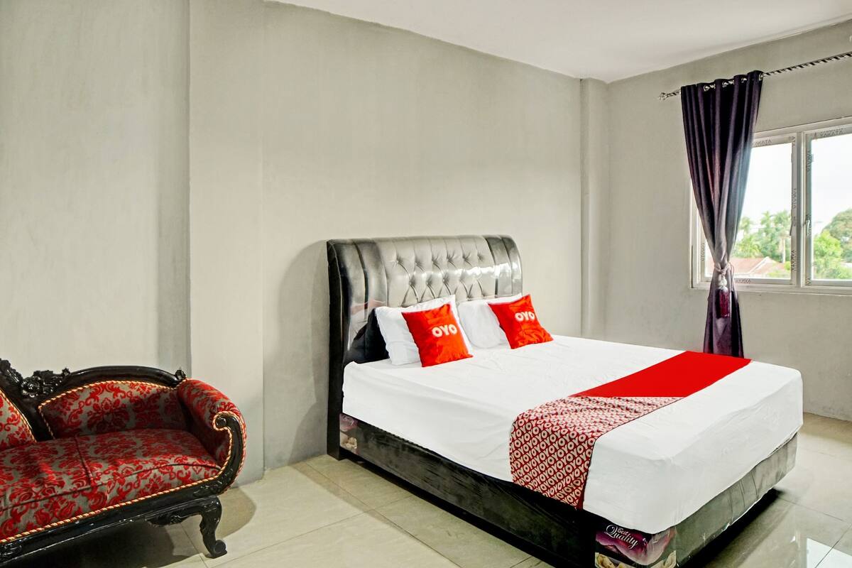 A bedroom with a large bed, a red and white patterned sofa, and a window with curtains.