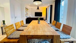 Een moderne woonkamer met een houten eettafel, omringd door banken en stoelen, een grote zwarte hanglamp en een flatscreen televisie aan de muur.
