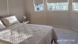 Quarto com cama, duas janelas, mesa de cabeceira e teto com vigas aparentes.