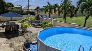 A swimming pool surrounded by lounge chairs and a blue umbrella, with palm trees and a house in the background. - Serene escape 2 bedroom with Pool (Oistins)