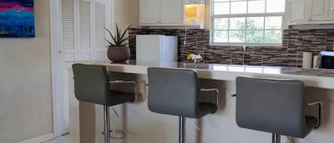 Una cocina con armarios blancos, una isla central con taburetes, una lámpara colgante y un ventanal que proporciona luz natural.