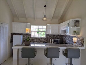 A kitchen with white cabinets, a stone backsplash, a central island with bar stools, and a hanging light fixture. - Serene escape 2 bedroom with Pool (Oistins)