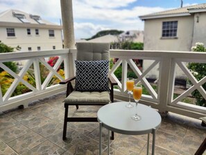 A balcony with a chair and a table, two glasses of orange juice, and a view of buildings in the background. - Serene escape 2 bedroom with Pool (Oistins)