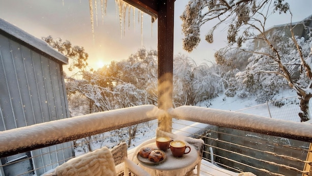 A balcony with a table set for two, a chair, and a view of snow-covered trees and a building. - Glacier Chalet - Luxury Mt Buller Ski Chalet • Fireplace •Sleeps 14 (Mount Buller)