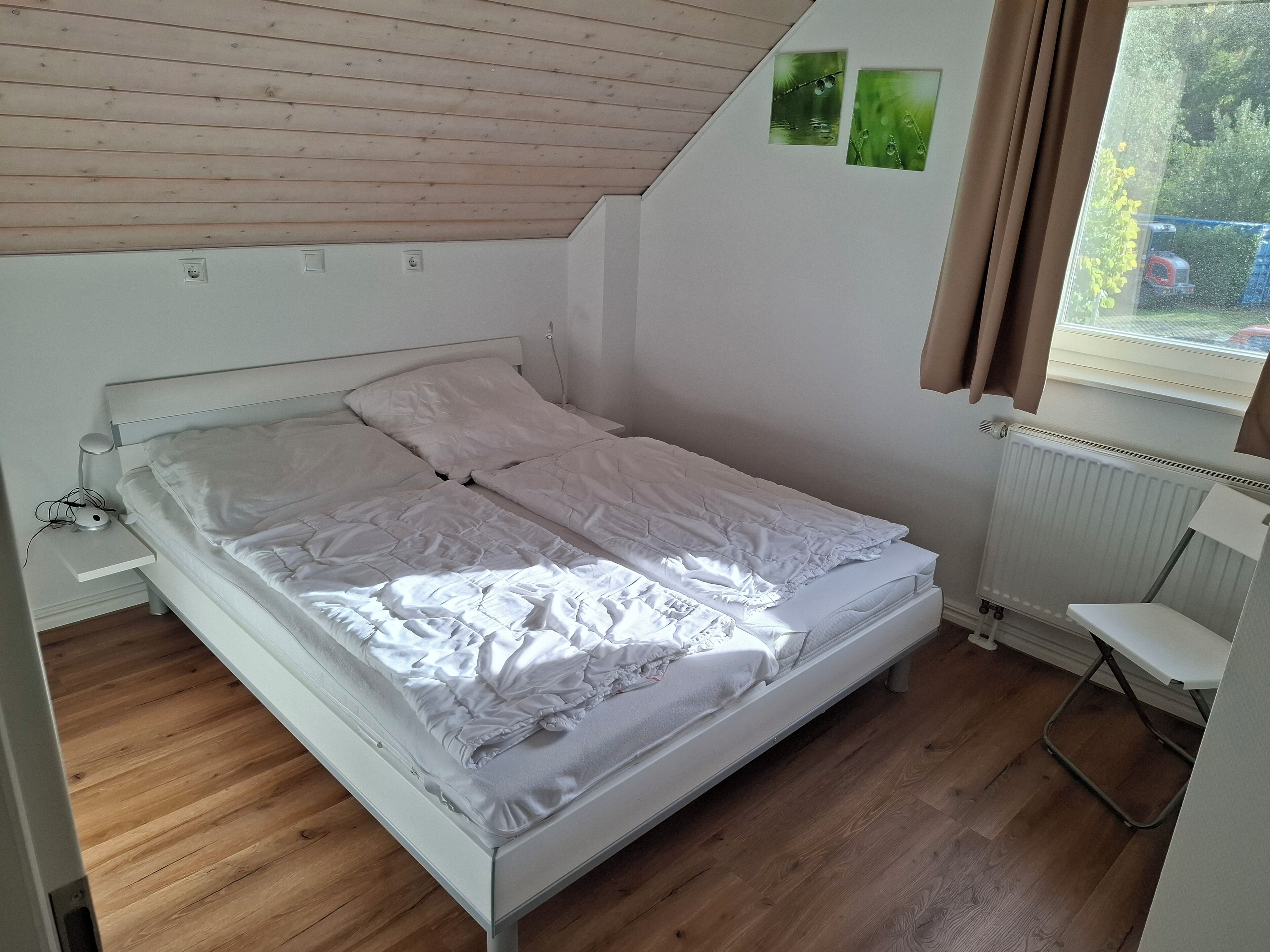 Een eenpersoonsbed met witte lakens in een kamer met houten vloeren, een witte radiator en een raam met gordijnen.